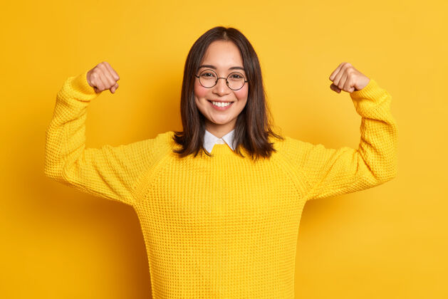活跃积极的亚洲年轻女子举手展示肌肉假装很强壮有力微笑着轻轻地穿上休闲毛衣态度女性眼镜