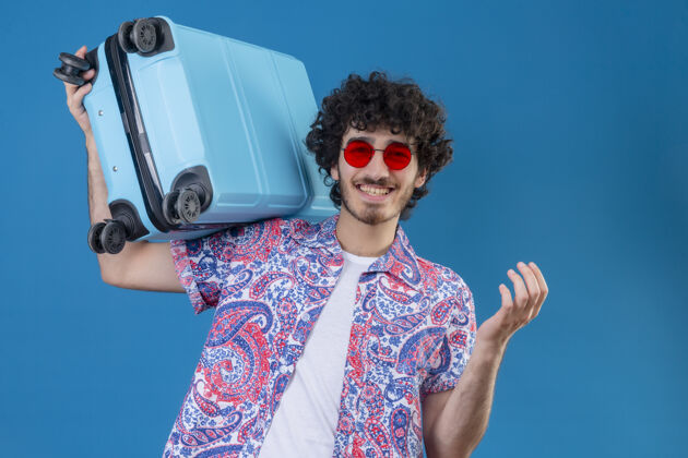 手提箱快乐的年轻英俊的卷发旅行者戴着太阳镜肩上提着手提箱在与世隔绝的蓝色空间与复制空间快乐蓝色帅气