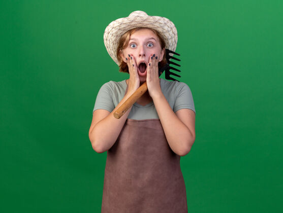 女性震惊的年轻斯拉夫女园丁戴着园艺帽把手放在脸上拿着绿色的耙子花园年轻人帽子