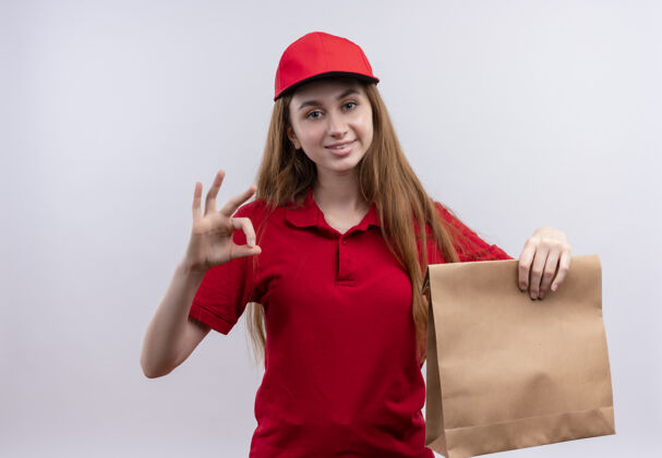 制服身着红色制服 面带微笑的年轻送货员 手拿纸袋 在与世隔绝的空白处做着“ok”的手势拿着红色女孩