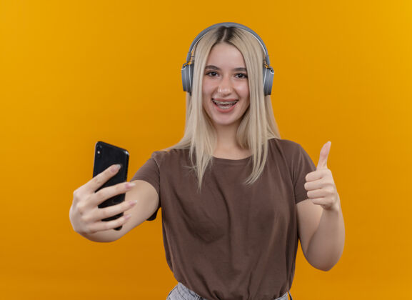 手机快乐的金发女孩戴着耳机戴着牙套拿着手机 在孤立的橙色空间里竖起大拇指拇指年轻电话