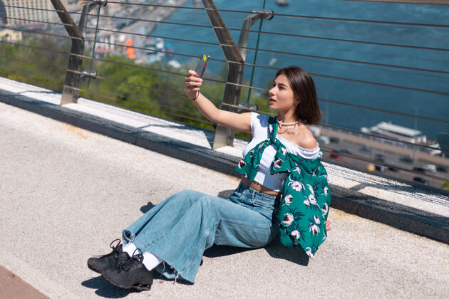 女性阳光明媚的日子里穿着休闲绿衬衫的女人站在桥上看手机屏幕的户外肖像自拍打视频电话应用表达成功