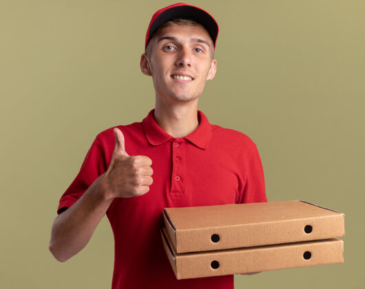 拇指微笑的年轻金发送货员竖起大拇指 拿着橄榄绿的比萨盒橄榄盒子金发