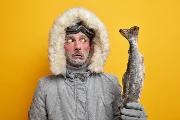 探险冬季钓鱼和运动概念惊呆了的冻人拿着惊呆了的大奖杯钓到的鱼穿的外套有着满脸积雪的红脸北方冰河
