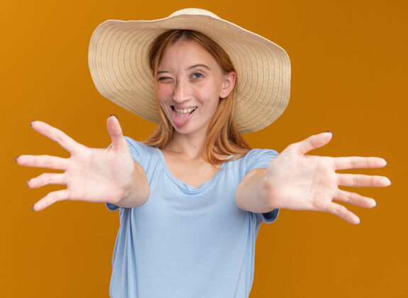 年轻快乐的年轻红发姜汁女孩 满脸雀斑 戴着沙滩帽 撅着舌头 眨着眼睛 伸出手来对着橘子姜快乐手