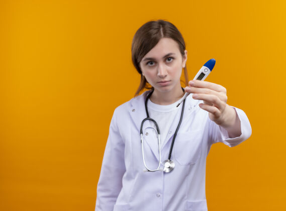 体温计严肃的年轻女医生穿着医用长袍和听诊器 在隔离的橙色空间和复印空间拉伸温度计医生医疗长袍