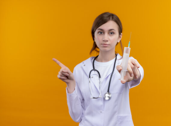 长袍严肃的年轻女医生穿着医用长袍和听诊器伸展注射器 指着左边的橙色空间和复印空间太空伸展听诊器
