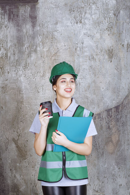 模特身着绿色制服 头戴安全帽的女工程师 手里拿着一个黑咖啡杯和一个蓝色项目文件夹检查表年轻人聪明