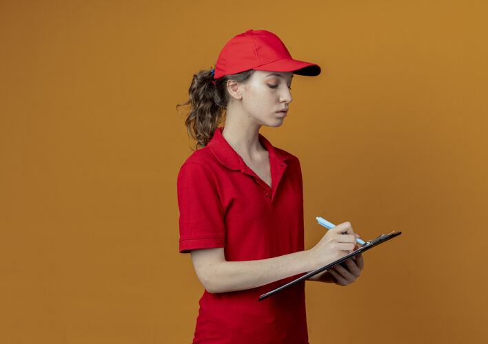 写字专注的年轻漂亮的送货女孩 穿着红色制服 戴着帽子 在剪贴板上写字 笔被隔离在橙色背景上 留有复印空间橙色剪贴板钢笔