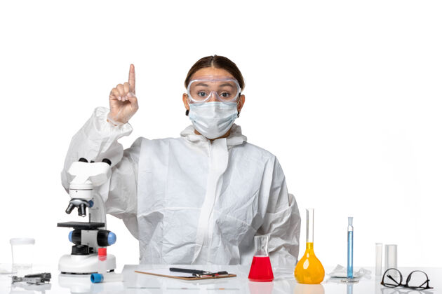 坐着正面图女医生穿着防护服 戴着口罩 坐在白色背景病毒的溶液里-健康大流行病毒实验室外套前面