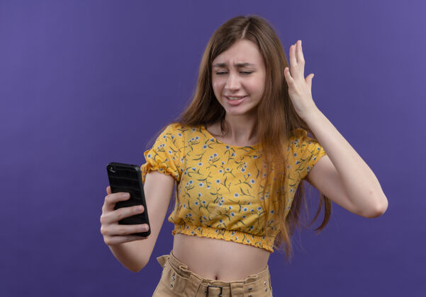 恼火恼怒的年轻女孩拿着手机 在孤立的紫色空间举手手年轻电话