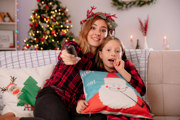 电视带着冬青花环的母亲手持电视遥控器 看着摄像机 女儿坐在沙发上 在家里享受圣诞节时光花圈印象时间