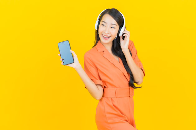 收音机肖像美丽的亚洲年轻女子戴着耳机和智能手机听黄色音乐橙色连身裤黄色肖像