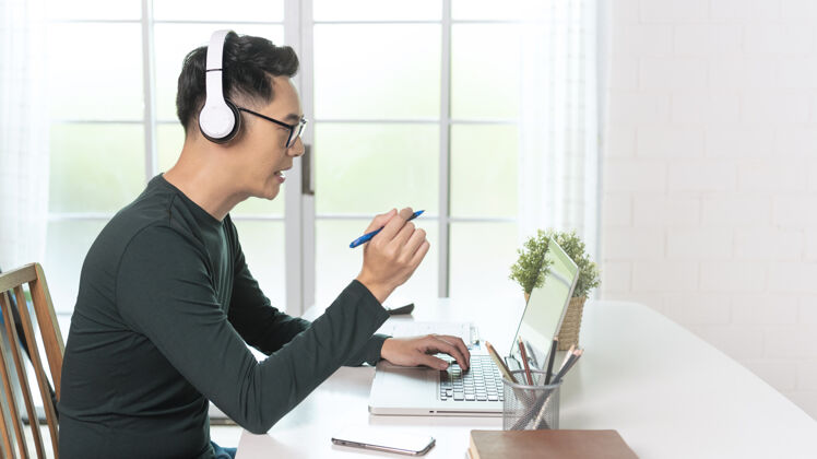 电脑微笑英俊的亚洲商人戴着耳机在家里远程工作他是网络研讨会视频会议互联网人办公桌