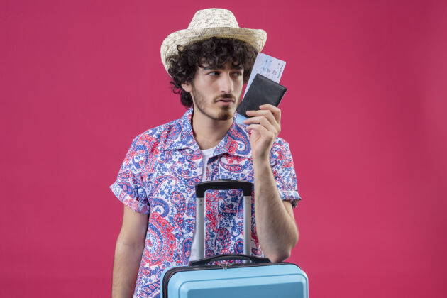 钱包自信的年轻帅气的卷发旅行者戴着帽子拿着钱包和机票 把胳膊放在手提箱上看着右边的粉色空间和复印空间持有帅气帽子