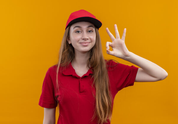 女孩穿着红色制服的自信的年轻送货女孩在孤立的橙色空间上做着ok标志橙色年轻制服