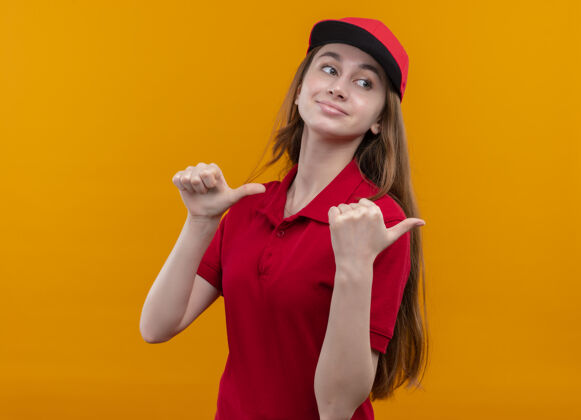 交付自信的年轻送货女孩穿着红色制服 右手竖起大拇指放在橙色的空白处拇指女孩橙色