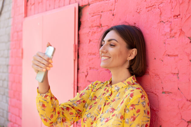 手机穿着黄色夏装的时髦女人在粉红色的砖墙上快乐地积极地在手机上自拍电话肖像网络