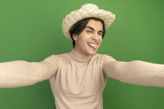 表情快乐的年轻帅哥戴着帽子露着舌头站在绿色的墙上快乐脸人