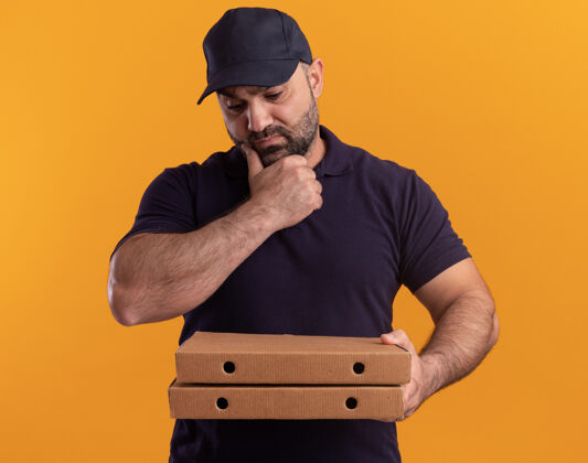 人想着中年送货员 穿着制服 戴着帽子 看着披萨盒 把下巴孤立在黄色的墙上表情站着盒子
