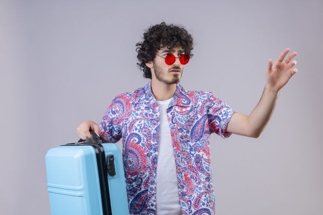 手困惑的年轻英俊的卷发旅行者戴着太阳镜拿着手提箱 伸出手在右侧孤立的白色空间手提箱帅气穿