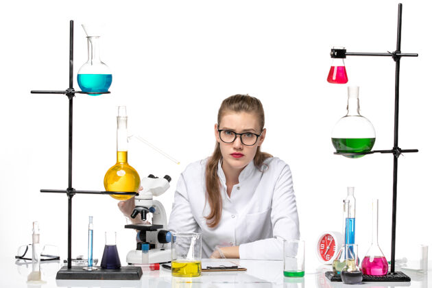 视野前视图穿着医疗服的女化学家坐在白色背景上 拿着溶液和显微镜大流行化学冠状病毒化学正面女化学家