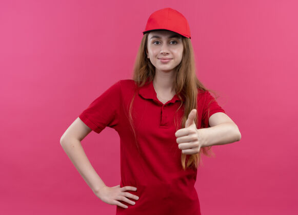 年轻身着红色制服的自信的年轻送货女孩在孤立的粉色空间里竖起大拇指 把手放在腰上向上粉色红色