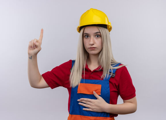 手令人印象深刻的年轻金发工程师建设者女孩在制服上指着与孤立的白色空间腹部手年轻女孩印象