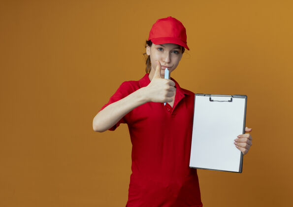 拇指自信的年轻漂亮的送货女孩 穿着红色制服 戴着帽子 手里拿着笔和剪贴板 对着橙色背景上的相机竖起大拇指 还有复印空间自信红色钢笔