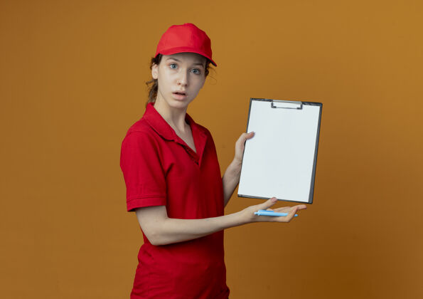 交货令人印象深刻的年轻漂亮的送货女孩在红色制服和帽子拿着钢笔和显示剪贴板上的橙色背景与复制空间隔离剪贴板背景帽子