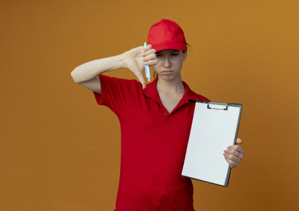 年轻未出租的年轻漂亮的送货女孩在红色制服和帽子举行钢笔和剪贴板显示拇指向下孤立的橙色背景与复印空间显示红色背景