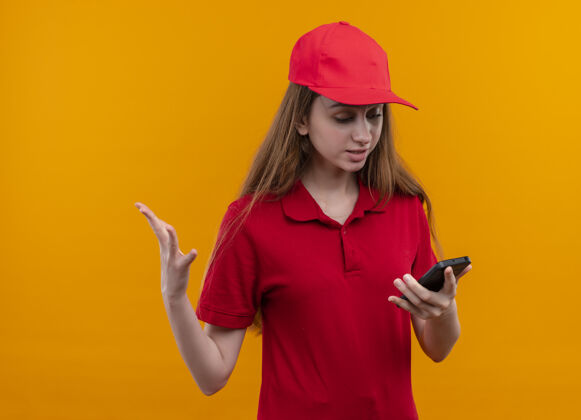橙色恼怒的年轻送货女孩穿着红色制服拿着手机在孤立的橙色空间看着它电话拿着制服