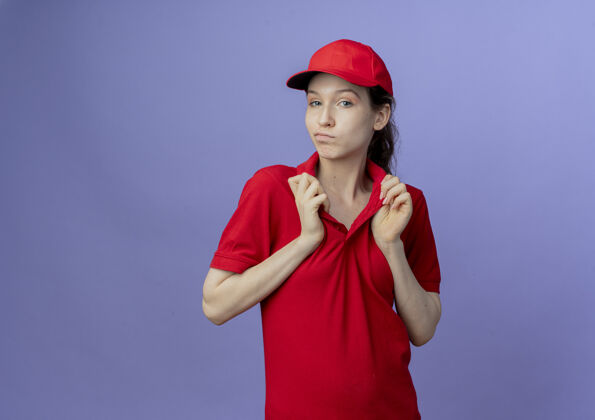自信自信的年轻漂亮的送货女孩穿着红色制服和帽子抓领她的t恤隔离紫色背景与复制空间制服背景衣领