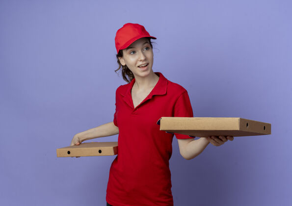 年轻令人印象深刻的年轻漂亮的送货女孩穿着红色制服和帽子举行比萨饼包看着一边孤立的紫色背景盒子手持帽子