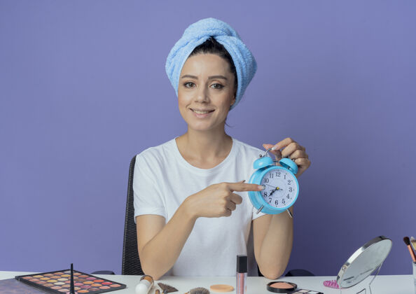 拿着微笑的年轻漂亮女孩坐在化妆桌旁 带着化妆工具 头上拿着浴巾 拿着并指着紫色背景上的闹钟坐着年轻漂亮