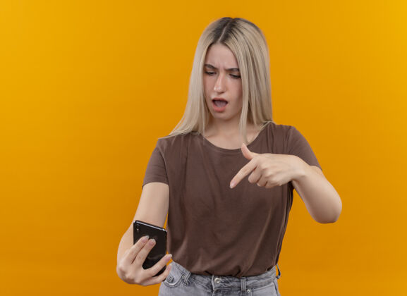 橙色困惑的年轻金发女孩拿着手机指着和看着它在孤立的橙色空间与复制空间金发手机电话