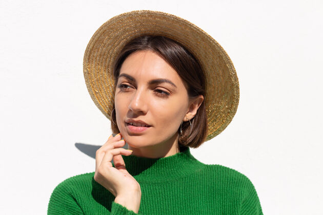 模型穿着绿色休闲毛衣和帽子的时尚女士户外白墙上平静自信的微笑墙脸阳光