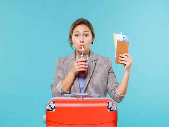 玻璃正面图：度假中的女性手持一杯新鲜的果汁和一张蓝色背景的机票 在海上度假旅行饮用航行