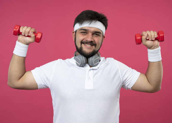 年轻微笑着的年轻人戴着头带和腕带 戴着耳机 用哑铃锻炼身体男人运动微笑