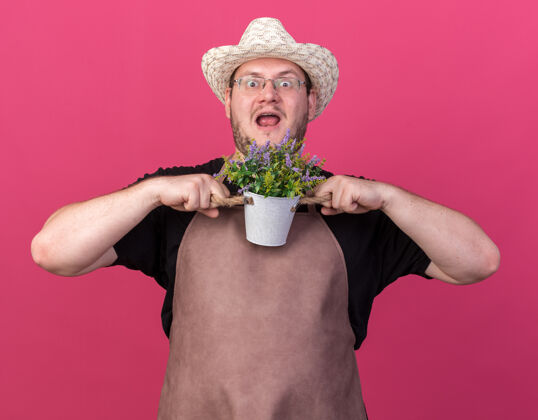 园艺惊讶的年轻男园丁戴着园艺帽 在粉红色的墙上孤立的花盆里捧着花市民人人