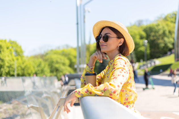 人穿着黄色夏装 戴着黄色帽子 喝着咖啡 享受阳光的女人的户外肖像 站在桥上 城市的景色令人惊叹20多岁城市景观茶