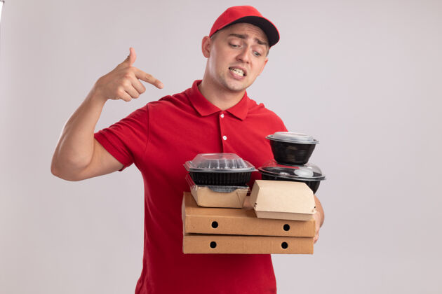 人困惑的年轻送货员穿着制服 戴着帽子 指着隔离在白墙上的比萨饼盒上的食品容器穿着食物制服