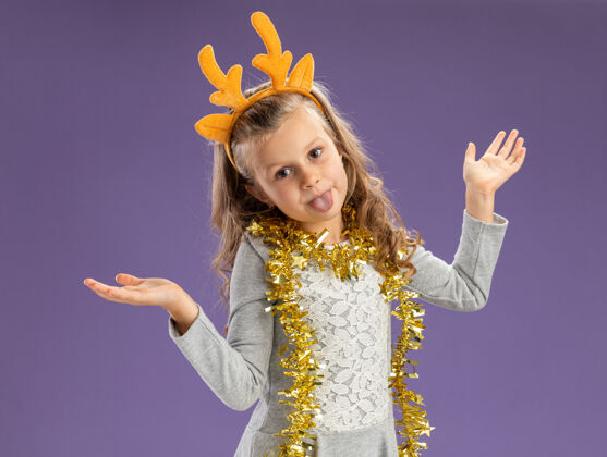 脖子快乐的小女孩戴着圣诞发箍 脖子上戴着花环 双手张开 在蓝色背景上露出孤立的舌头女孩箍圣诞快乐