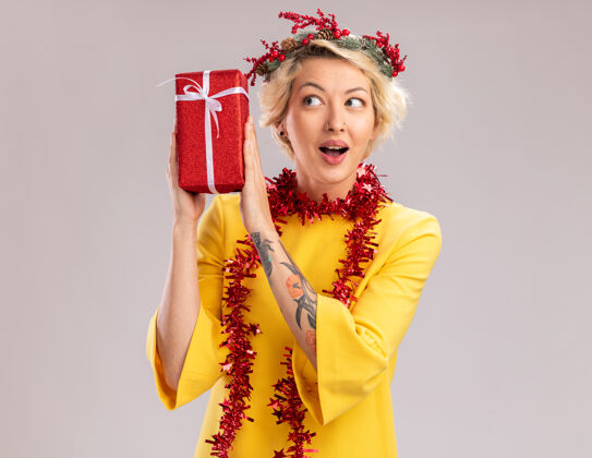 包装好奇的金发女郎头戴圣诞花环 脖子上戴着金箔花环 头旁拿着圣诞礼包 在白色背景下看着它头年轻空间