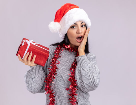 包装关心的年轻白人女孩戴着圣诞帽 脖子上戴着金箔花环 看着相机 手里拿着礼包 手放在脸上 在白色的背景下与世隔绝周围脸白色