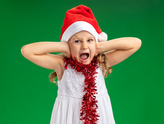 小恼怒的小女孩戴着圣诞帽 脖子上戴着花环 耳朵被隔离在绿色背景上圣诞快乐圣诞节女孩