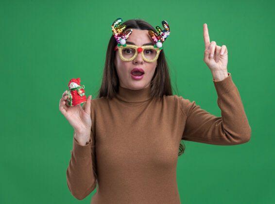 圣诞节印象深刻的年轻漂亮女孩穿着棕色毛衣和圣诞眼镜举行圣诞玩具点在绿色背景上孤立向上印象眼镜