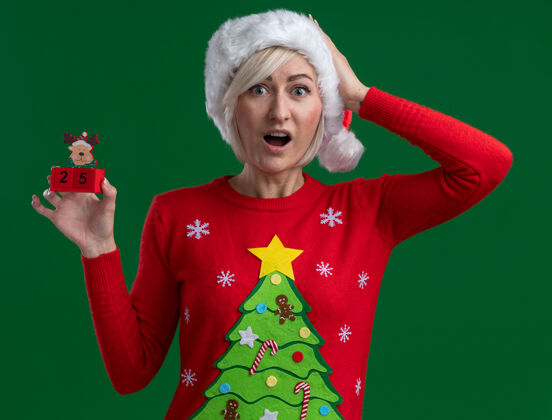 圣诞节令人印象深刻的中年金发女人戴着圣诞帽和毛衣拿着圣诞驯鹿玩具和日期看着相机保持手在头上隔离绿色背景举行头印象