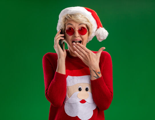 印象令人印象深刻的年轻金发女人戴着圣诞帽和圣诞老人的圣诞毛衣戴着眼镜讲电话看着相机保持手放在嘴上绿色背景隔离嘴圣诞老人帽子