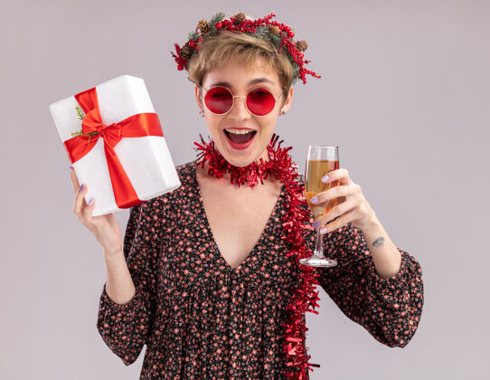 年轻快乐的年轻漂亮女孩戴着圣诞花环 脖子上戴着金箔花环 戴着眼镜 手里拿着礼包 手里拿着一杯香槟 看着相机在白色背景上眨眼花环头眼镜